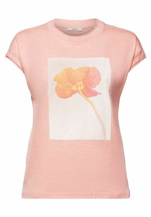 Persoon die een lichtroze T-shirt draagt met een grote oranje bloemenprint en witte broek met hoge taille, één hand in de zak, kettingketting.