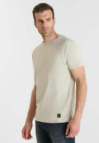 Uomo che indossa una semplice t-shirt beige a maniche corte e jeans scuri, in piedi contro uno sfondo bianco, che guarda a sinistra.