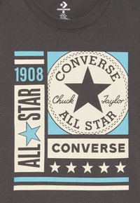Converse All Star-logo met een grote ster, de tekst "1908" en "Chuck Taylor" in blauw, wit en zwart op een donkere stof achtergrond.