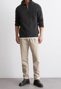 Dunkelgrauer, gerippter Strickpullover mit einem Viertel-Reißverschlusskragen, getragen über einem weißen Hemd; kombiniert mit beigen Hosen und schwarzen Knöchelstiefeln.