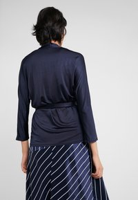 Námořnický modrý top s dlouhým rukávem se hladkým, lesklým povrchem a páskem v pase, spárovaný s pruhovanou sukní v námořnické modré a bílé.