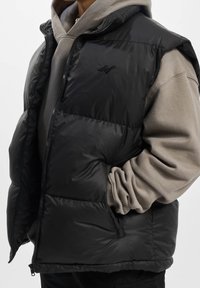 Czarna kamizelka puchowa z zamkiem błyskawicznym i bocznymi kieszeniami, nałożona na jasnoszary hoodie. Gładka tekstura o lekkiej konstrukcji.