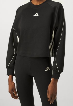 Osoba ubrana w czarną krótką bluzę Adidas z długim rękawem oraz czarne legginsy z białymi paskami i logo Adidas na piersi i udzie.