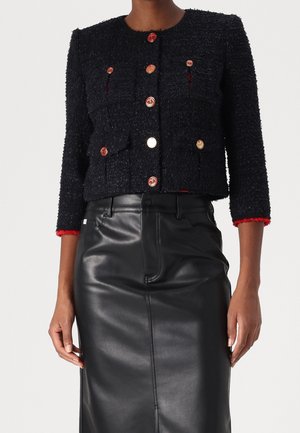 Mujer con una chaqueta corta negra texturizada con botones dorados y ribete rojo en los puños, combinada con una falda negra de cuero de talle alto.