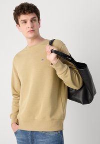 Khaki crewneck trui met geribde manchetten en zoom, met een klein geborduurd logo op de borst. Zwarte leren handtas die met één hand wordt vastgehouden.