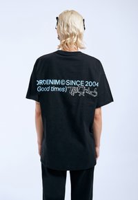 Černé oversized tričko vyrobené z bavlny. Na zadní straně se nachází modrý text "ORDENIM© SINCE 2004" a grafické prvky. Jednoduchý design, krátké rukávy.