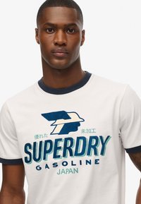 Camisa de algodón blanca con cuello de canalé azul marino, que presenta un gran gráfico con "SUPERDRY" en azul y verde, acompañado de texto en japonés.
