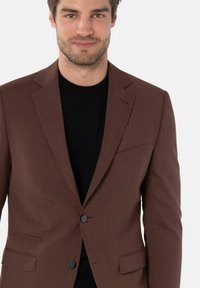 Brauner Blazer aus strukturiertem Stoff, mit einem Knopfverschluss und seitlichen Taschen, über einem schwarzen Pullover getragen.