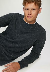 Donkergrijze trui met een gestructureerde afwerking, ronde halslijn en geribde manchetten. Model poseert casual en toont de ontspannen pasvorm van de trui.