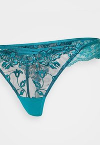 Τανγκάκι σε τιρκουάζ δαντέλα με λουλουδάτη κεντητική, διάφανα πάνελ και απαλό σατινέ λάστιχο. Οι υφασμάτινες άκρες προσθέτουν στην σχεδίαση του.