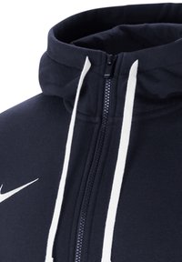 Marinblå zip-up hoodie tillverkad av mjukt tyg, med hög krage, vita dragband och en liten vit logo på vänster bröst.