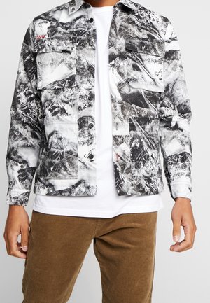 Homme portant une veste à motif abstrait noir et blanc sur une chemise blanche avec un pantalon en velours côtelé marron, les bras relâchés le long du corps.