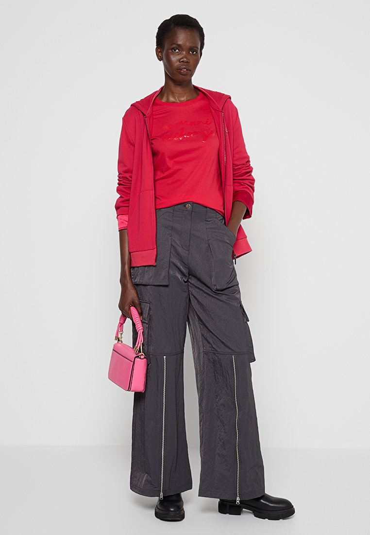 Donna che indossa un felpa rossa e una t-shirt, pantaloni cargo grigi a gamba larga con zip, stivali neri, mentre tiene una piccola borsa rosa.