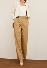 Cardigan beige côtelé porté sur un chemisier blanc, associé à un pantalon taille haute kaki avec une ceinture à ferrures argentées et des poches.