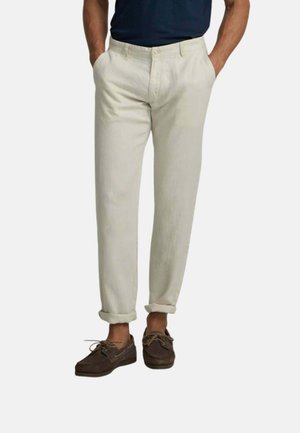 Hombre con pantalones beige remangados, camisa azul marino y zapatos náuticos marrones, de pie con las manos en los bolsillos.