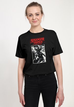 Kvinde med lys hud og blondt hår iført sort Stranger Things T-shirt med rød tekst og sort-hvidt monsterbillede.