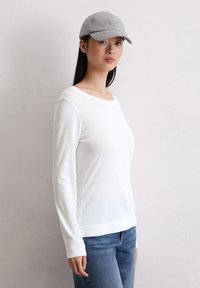 Camicia bianca a maniche lunghe con scollatura ampia, abbinata a jeans blu. Un berretto grigio con logo completa l'outfit. Design semplice e casual.