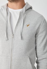 Sudadera con cremallera gris hecha de una tela suave. Presenta una cremallera frontal, dos bolsillos y un pequeño logo de un pájaro dorado en el lado izquierdo del pecho.