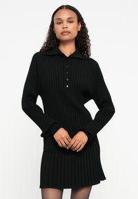 Abito in maglia nera a coste con colletto stile polo, placket con cinque bottoni e maniche lunghe. Texture morbida e silhouette aderente.