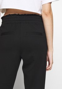 Svarta högmidjade byxor med en rynkad elastisk midja, som har två bakfickor och en slät textur. Designen är skräddarsydd och åtsittande.