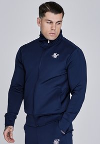 Mornarska zip-up jakna izrađena od glatkog materijala, s visokim ovratnikom, bočnim džepovima i bijelim logotipom na prsima. Uz tijelo prilagođen dizajn.