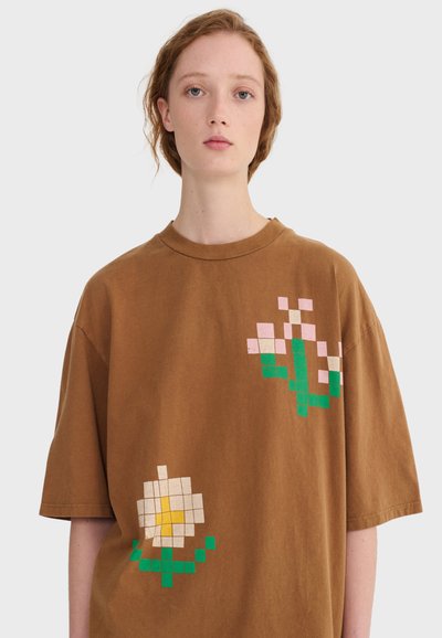 Mlada osoba svijetle kože i kestenjaste kose nosi smeđu preveliku majicu s pikseliziranim dizajnom cvijeća na bijeloj pozadini.