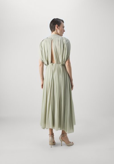 Rochie verde deschis cu textură pliată și detalii cu spatele deschis, având mâneci scurte și talie ajustată. Modelul poartă sandale bej cu toc înalt.