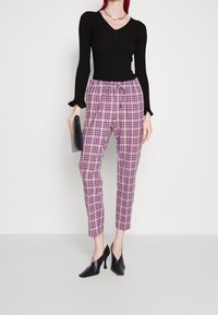 Schwarzes, geripptes Langarmoberteil, karierte Hose mit violetten, rosa und beige Farbtönen, elastischem Bund und schwarzen spitzen Absätzen. Hält eine schwarze Clutch.