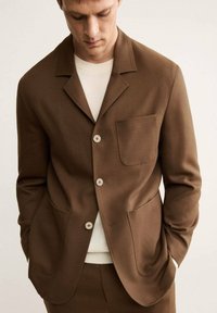 Blazer marrone su misura in tessuto liscio, con colletto a risvolto, tasca singola e chiusura a tre bottoni. Abbinato a una camicia bianca a girocollo.