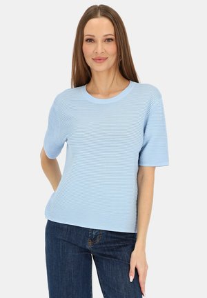 Kobieta z prostymi brązowymi włosami, ubrana w jasnoniebieską prążkowaną bluzkę z krótkim rękawem i ciemnoniebieskie jeansy, stojąca z jedną ręką za plecami.