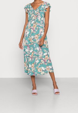 Robe midi turquoise à imprimé floral avec décolleté en V à volants et manches courtes, portée avec des sandales à talons ouvertes violet clair sur fond uni.