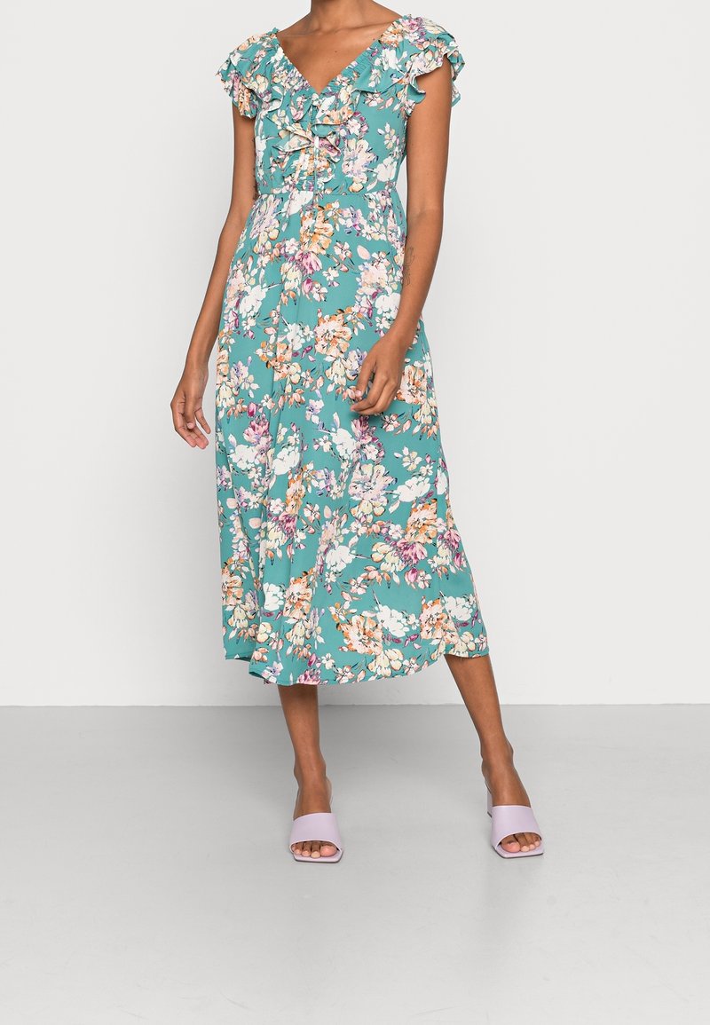 Robe midi turquoise à imprimé floral avec décolleté en V à volants et manches courtes, portée avec des sandales à talons ouvertes violet clair sur fond uni.