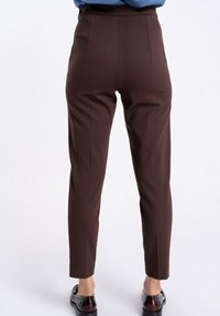 Osoba ubrana w brązowe spodnie o wysokim stanie i zwężane nogawki oraz ciemne, błyszczące półbuty stojąca na tle jednolitej białej ściany.