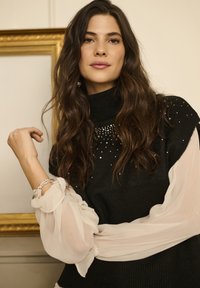Črn pulover z visokim ovratnikom in okrašenimi kroglicami, združen s prosojnimi belimi rokavi. Modelka pozira z zapestnico na zapestju.