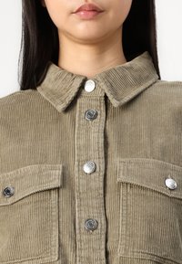 Chemise en velours côtelé de couleur olive, avec un col, des boutons-pression en métal argenté et des poches poitrine à texture nervurée.