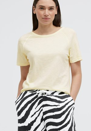 Frau trägt ein hellgelbes kurzärmeliges Hemd und schwarz-weiß gestreifte Zebrahosen, Hände in den Taschen, vor einem einfarbigen Hintergrund.