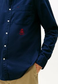 Marine katoen overhemd met knopen en een zak, voorzien van een rood geborduurd logo en witte knopen; gladde textuur en stof van gemiddelde dikte.