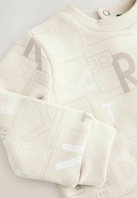 Jasno beżowy sweatshirt o teksturowanym wykończeniu, z subtelnym białym wzorem alfabetu. Okrągły dekolt i ściągacze przy rękawach.