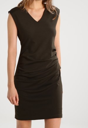 Tmavě hnědé šaty bez rukávů do kolen s výstřihem do V a nařaseným detailem v pase, nošené stojící osobou.
