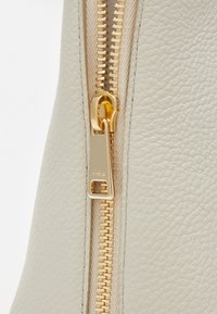 Dettaglio di una borsa in pelle color crema con una texture liscia, cerniera dorata e una targhetta del marchio con la scritta "Furla" in rilievo.