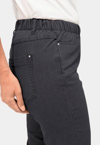 Donkergrijze elastische broek met een getextureerde stof. Voorzien van twee achterkantzakken en een soepele zijnaad, getoond vanuit een zijaanzicht.