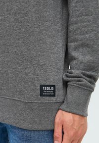 Nærbilde av en persons hånd og underarm iført en grå hettegenser med en svart etikett som sier "!SOLID For hverdagens ro."