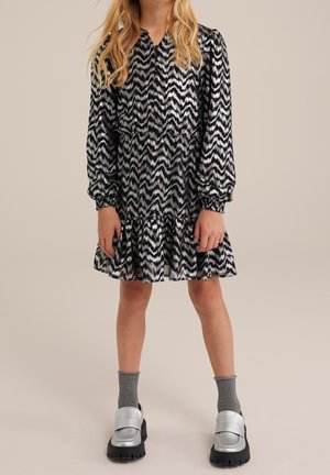 Dziecko ubrane w długą sukienkę w czarno-biały wzór w zygzak, szare skarpetki i srebrne buty na grubym spodzie na jednolitym tle.