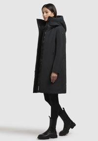 Long manteau en duvet noir avec un col haut, des poches zippées sur les côtés et une texture lisse. Porté sur des leggings noirs et des bottes noires épaisses.