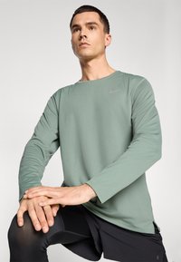Chemise à manches longues en tissu vert muted au toucher lisse; présente un col rond et un logo Nike subtil en gris.