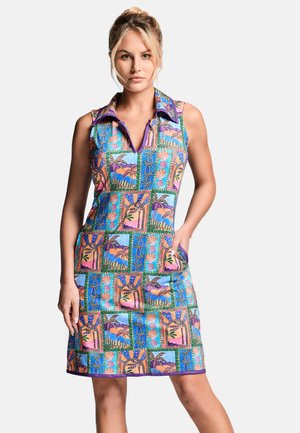 Kobieta ubrana w sukienkę bez rękawów z kołnierzykiem, z kolorowym tropikalnym wzorem przedstawiającym palmy i abstrakcyjne wzory, stoi z jedną ręką w kieszeni.