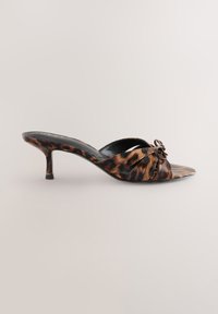 Mules à talons imprimé léopard avec un design à bride croisée, une texture lisse et un petit talon aiguille. Accentuées par un nœud décoratif.