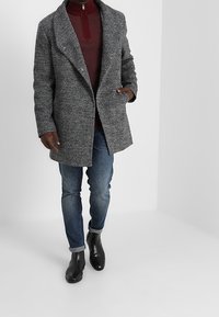 Grijze, gestructureerde wollen jas met een sjawlkraag, gecombineerd met een bordeauxrode ritstas en blauwe jeans. Zwarte enkelboots maken de look compleet.