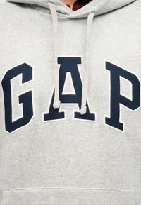 Sweat à capuche gris clair avec les lettres "GAP" en bleu marine sur la poitrine et des cordons suspendus à la capuche.