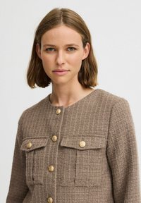 Chaqueta marrón texturizada con un patrón tejido, que presenta botones dorados y dos bolsillos en el pecho. Diseño abotonado con un escote redondo.